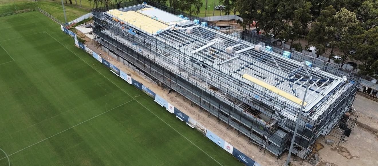Aerial view of a two-storey sports pavilion under scaffold with steel roof framing exposed
